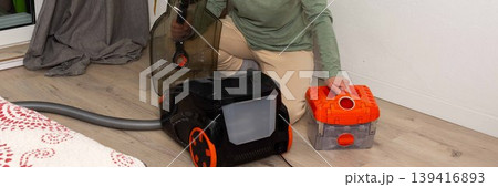 A person is getting ready to vacuum a room. They are attaching a container to the vacuum cleaner while kneeling on the floor. A plant is visible in the background, banner A person is getting ready to vacuum a room. They are attaching a container to the vacuum cleaner while kneeling on the floor. A plant is visible in the background, banner 139416893