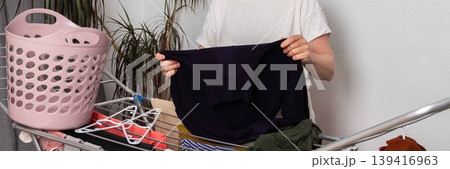 A 50-year-old woman is in her room. She is hanging washed laundry on a drying rack. A basket of clothes is nearby. She looks focused on her task, banner A 50-year-old woman is in her room. She is hanging washed laundry on a drying rack. A basket of clothes is nearby. She looks focused on her task, banner 139416963