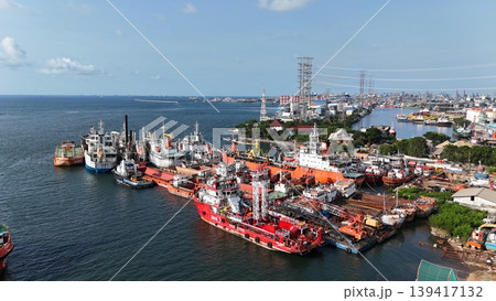 A red offshore supply and rescue ship equipped with a lattice tower, docked at a busy industrial port alongside various barges and heavy maritime equipment. 139417132