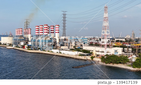 Aerial view of a coastal natural gas power plant station with striped industrial chimneys, high voltage transmission towers, and electrical infrastructure against a clear blue sky. Aerial view of a coastal natural gas power plant station with striped industrial chimneys, high voltage transmission towers, and electrical infrastructure against a clear blue sky. 139417139