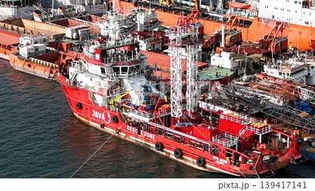 A red offshore supply and rescue ship equipped with a lattice tower, docked at a busy industrial port alongside various barges and heavy maritime equipment. 139417141
