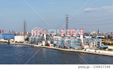 Aerial view of a coastal natural gas power plant station with striped industrial chimneys, high voltage transmission towers, and electrical infrastructure against a clear blue sky. Aerial view of a coastal natural gas power plant station with striped industrial chimneys, high voltage transmission towers, and electrical infrastructure against a clear blue sky. 139417149