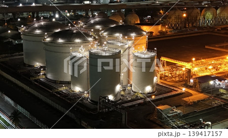 High-angle night view of a fuel storage tank terminal with multiple large, illuminated cylinders and a complex piping infrastructure at an industrial port facility, glowing in the dark. 139417157