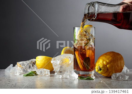 Iced black tea is poured from a bottle into a tall glass with lemon wedges, ice cubes and mint leaves. 139417461