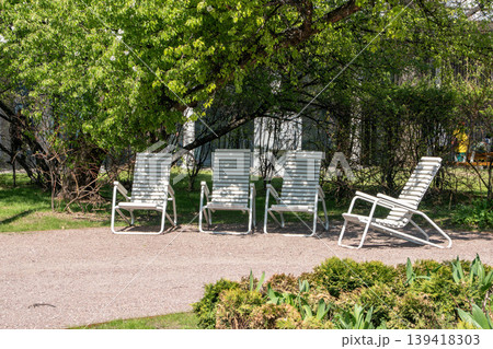 White metal lounge chairs stand on gravel in front of blooming tree with dense white flowers, green foliage in spring park. Outdoor furniture, seasonal bloom, relaxation, urban greenery, lifestyle 139418303