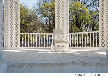 White lounge chair stands on terrace under decorative pavilion with columns and lattice structure overlooking trees. Outdoor furniture, architectural detail, relaxation space, garden design, lifestyle 139418306