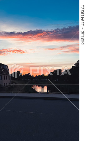 Serene sunset over calm river with vibrant colors reflecting on water, framed by trees and buildings, creating a tranquil evening atmosphere Serene sunset over calm river with vibrant colors reflecting on water, framed by trees and buildings, creating a tranquil evening atmosphere 139419221