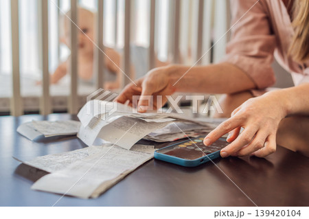 Mother sitting on sofa calculating monthly expenses with receipts while baby lies in crib nearby at home. Stressed mom managing budget and childcare. Financial pressure and parenthood concept Mother sitting on sofa calculating monthly expenses with receipts while baby lies in crib nearby at home. Stressed mom managing budget and childcare. Financial pressure and parenthood concept 139420104