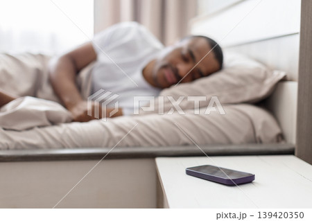 Smartphone on bedside table with black man sleeping in background in bedroom Smartphone on bedside table with black man sleeping in background in bedroom 139420350