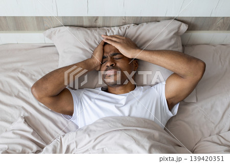 Black man lying in bed holding head with pain expression in bedroom 139420351
