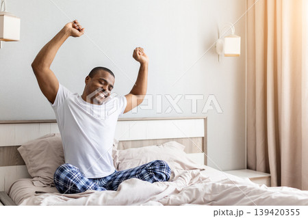 African american man stretching arms while sitting in bed in bedroom 139420355