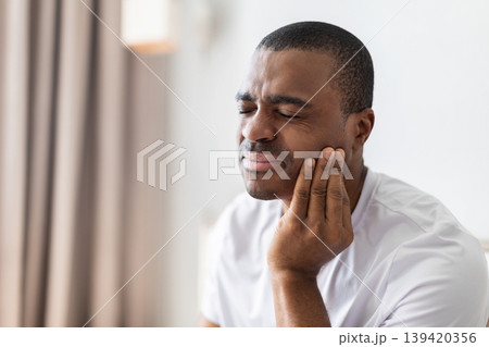 African american man sitting on bed touching cheek with pain expression 139420356