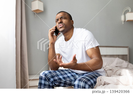 Black man talking on smartphone while sitting on bed, making expressive gesture 139420366