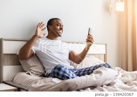 Happy black man sitting in bed, waving at smartphone during video call 139420367
