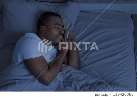 African american man sleeping at night in bed with hands under pillow African american man sleeping at night in bed with hands under pillow 139420369