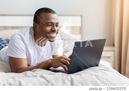 African american man lying on bed using laptop with smile in bedroom African american man lying on bed using laptop with smile in bedroom 139420370