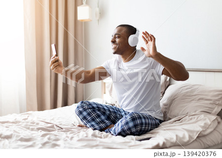 Smiling african american man wearing headphones waving at smartphone on bed Smiling african american man wearing headphones waving at smartphone on bed 139420376