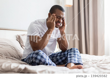 African american man sitting on bed holding temples with pain in bedroom African american man sitting on bed holding temples with pain in bedroom 139420382