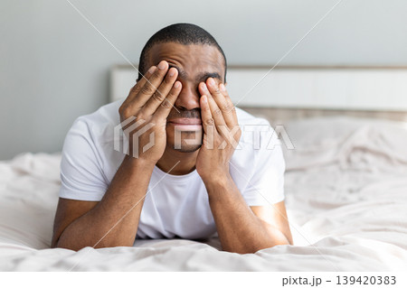 African american man lying on bed covering face with hands, expressing stress African american man lying on bed covering face with hands, expressing stress 139420383