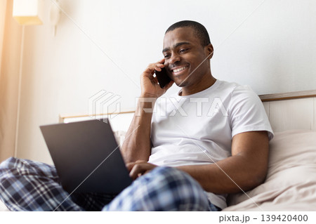 African american man talking on smartphone while using laptop on bed African american man talking on smartphone while using laptop on bed 139420400
