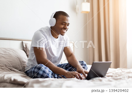 African american man sitting on bed typing on laptop in bedroom 139420402