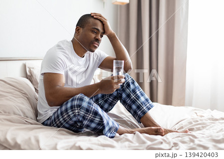 African american man sitting on bed holding glass of water, looking tired 139420403