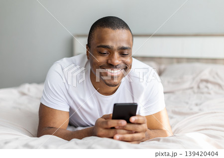 Smiling african american man lying on bed using smartphone in bright bedroom 139420404