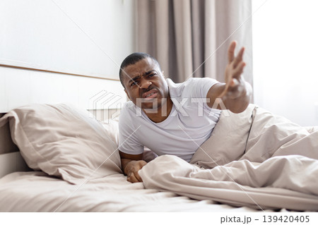 Frustrated black man lying in bed, reaching arm out with irritated expression 139420405