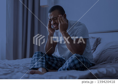 African american man sitting on bed with headache under blue night lighting African american man sitting on bed with headache under blue night lighting 139420418