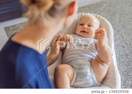 Mother talking to baby lying in cozy lounger on kitchen table while cooking at home. Caring mom interacting with infant during daily routine. Parenting, multitasking and family life concept 139420508