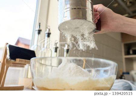 Hand sifting flour into glass bowl with batter on kitchen counter Hand sifting flour into glass bowl with batter on kitchen counter 139420515