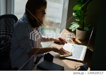 Woman wiping laptop keyboard with cloth while sitting at desk near window 139420517