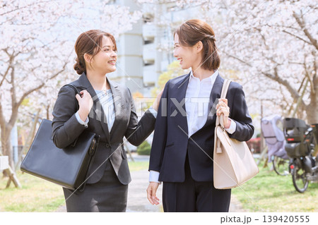 桜並木の下を歩きながら笑顔で会話する二人の若手女性ビジネスパーソン・春の通勤風景 139420555