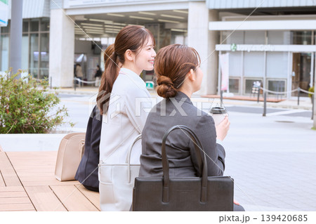 駅前のベンチに座って会話する二人の若い女性ビジネスパーソン・スーツ姿のOLがコーヒーを持ちながら休憩 139420685