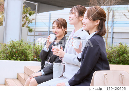 屋外のベンチでコーヒーを飲みながら休憩する3人の若い女性ビジネスパーソン・スーツ姿の新入社員 139420696