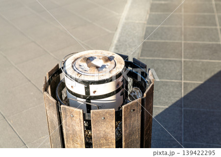 Traditional ceramic charcoal brazier discarded in a wooden public garbage bin on a city walkway. Disposed cooking stove in urban environment under sunlight. High angle view of refuse container. Traditional ceramic charcoal brazier discarded in a wooden public garbage bin on a city walkway. Disposed cooking stove in urban environment under sunlight. High angle view of refuse container. 139422295