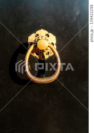 Ornate brass ring door knocker with a floral base plate mounted on a dark textured surface. Traditional Japanese architectural detail with golden hardware, bright sunlight, and dramatic shadows. Ornate brass ring door knocker with a floral base plate mounted on a dark textured surface. Traditional Japanese architectural detail with golden hardware, bright sunlight, and dramatic shadows. 139422298