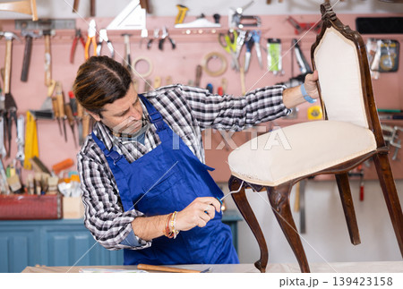 Middle-aged male restorer removes old cloth seat covering from antique wooden chair. 139423158