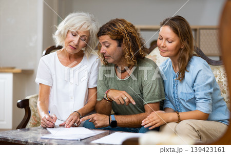 Elderly woman at table in home room filling up documents with family Elderly woman at table in home room filling up documents with family 139423161