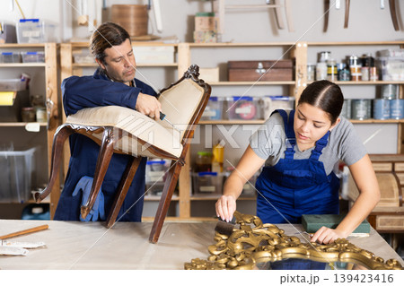 Skilled man and woman furniture restorers in uniform restoring old-fashioned chair mirror frame in workshop Skilled man and woman furniture restorers in uniform restoring old-fashioned chair mirror frame in workshop 139423416