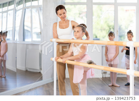 Girl ballet students prepare for classes, near ballet bare stretch their leg under teacher guidance. Girl ballet students prepare for classes, near ballet bare stretch their leg under teacher guidance. 139423753