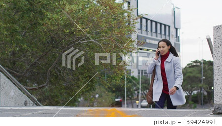 Asian woman in grey coat ascending arrow-marked steps in plaza, talking on smartphone, copy space 139424991