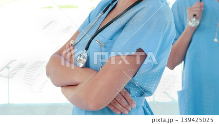 Standing nurse in scrubs crossing arms, colleague holding stethoscope in hospital hallway, sunlight Standing nurse in scrubs crossing arms, colleague holding stethoscope in hospital hallway, sunlight 139425025
