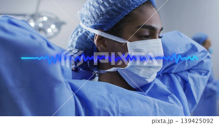 Surgeon adjusting mask straps in operating room, with blue scrubs, cap, lamp and ECG waveform Surgeon adjusting mask straps in operating room, with blue scrubs, cap, lamp and ECG waveform 139425092