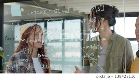 Women in varied attire collaborating on code behind glass board in office, with pastel sticky note Women in varied attire collaborating on code behind glass board in office, with pastel sticky note 139425110