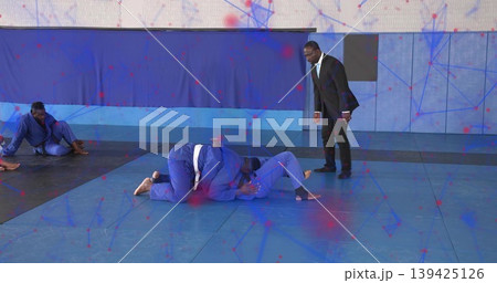 Grappling partners wearing blue gis, white belts on dojo padded mats, with coach in suit observing Grappling partners wearing blue gis, white belts on dojo padded mats, with coach in suit observing 139425126