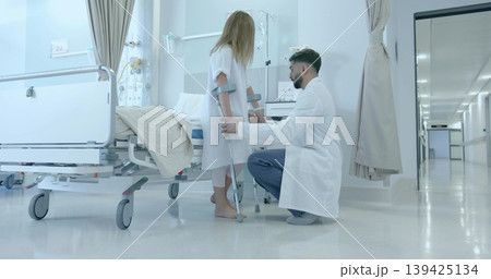 Kneeling doctor helping child in gown using crutches in ward, with bed, IV pole, cart, curtains 139425134