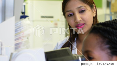 Mid adult Indian scientist in white lab coat guiding trainee at lab bench, with compound microscope Mid adult Indian scientist in white lab coat guiding trainee at lab bench, with compound microscope 139425236