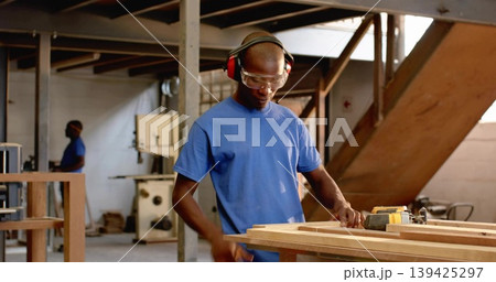 Sanding carpenter wearing goggles and earmuffs using power sander at workshop, wooden planks 139425297