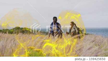 Hiking couple wearing jackets walking through tall dune grass by ocean, with packs poles sleep pad Hiking couple wearing jackets walking through tall dune grass by ocean, with packs poles sleep pad 139425650